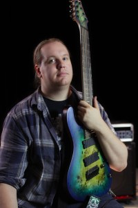 Man looks down the camera lens, holding a green, 8-string electric guitar
