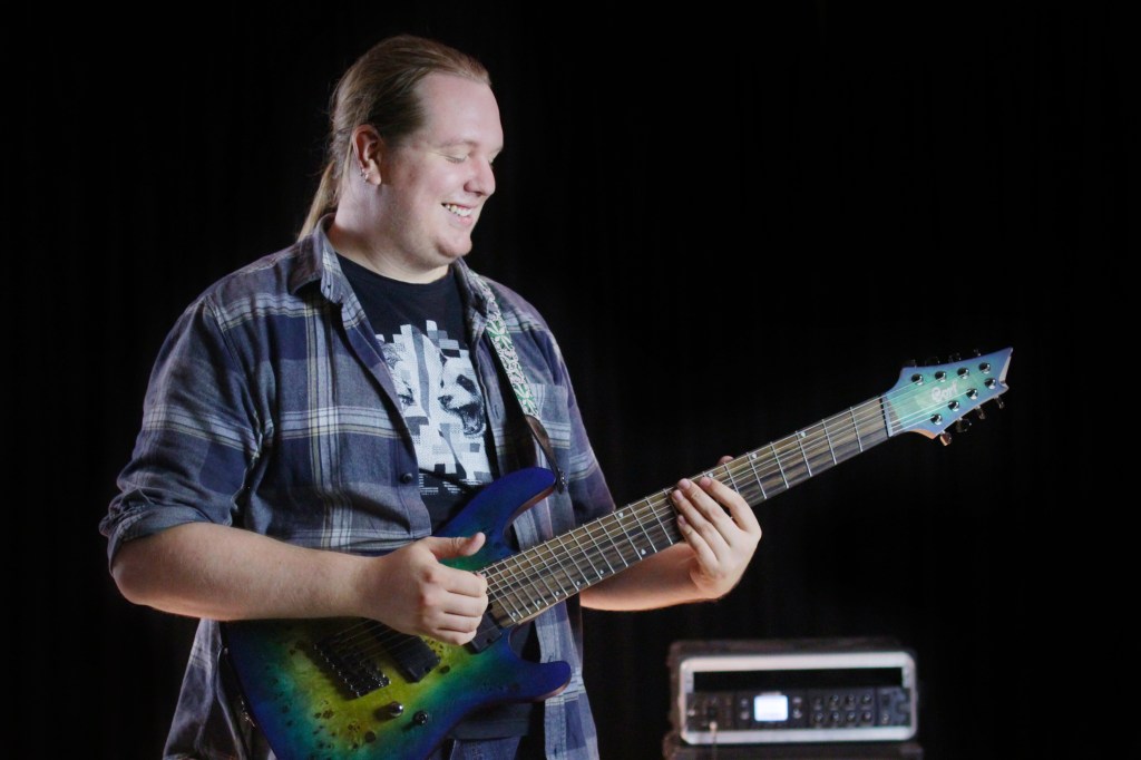 Man smiles whilst playing a green 8-string electric guitar, fingerstyle.